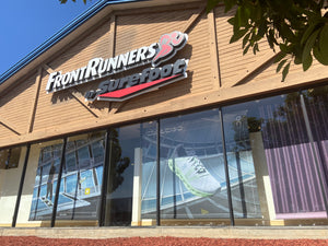 A photo of the front of the Front Runners LA Store in South Bay. A wood building with the sign reading FrontRunners in white and by Surefoot underneath in Black. Large windows display window decals for the brand On