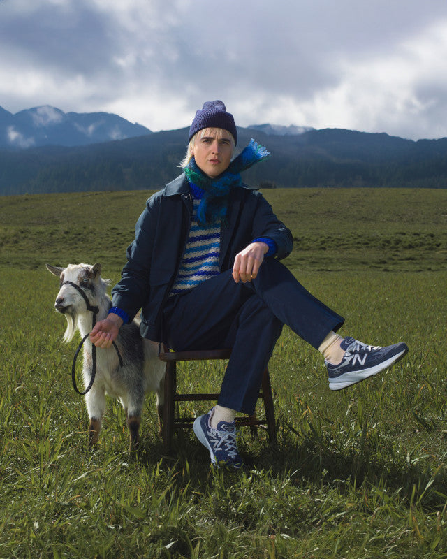 This picture is hilarious.  It shows a young man wearing 990's sitting in a green grass field with a goat