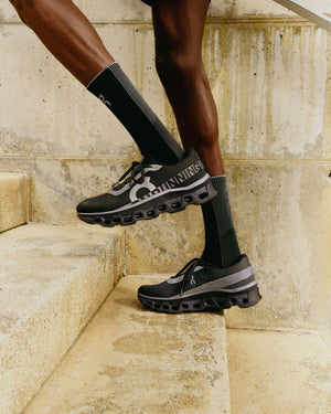 Up close view of the left side of a male from the knee down walking up stairs wearing black socks and footwear from the brand On against a neutral background color.