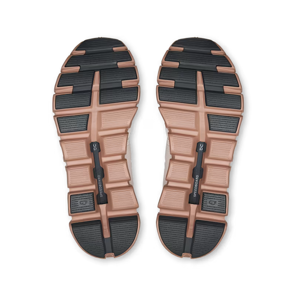 A pair of shoes shown from the bottom, displaying their rugged outsoles. The soles feature a dual-tone design with a dusty rose/mauve midsole and black rubber tread blocks.