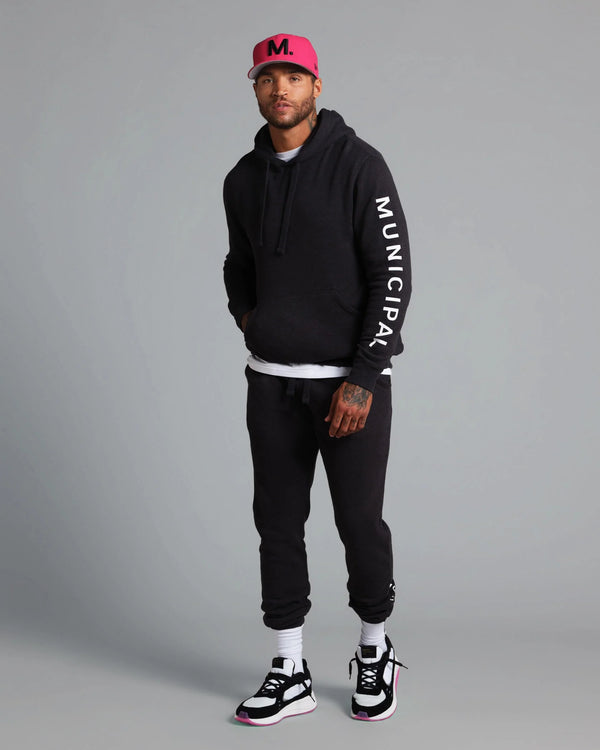 Man wearing a black hoodie with 'Municipal' text, black pants, and a pink cap on a gray background