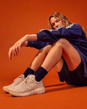 A female with blond hair sitting down with her knees up and arms resting on her knees wearing a blue jacket, black shorts and socks and pink VEJA footwear against an orange background