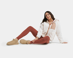 A women with long brown hair sitting down with her legs out front and to the side wearing beige ankle high UGG boots, red leggings and a white and beige fleece