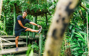 A man in a tropical jungle setting on a rustic deck stretching his left leg that is elevated on a railing.  His shirt and shorts are black and he's in focus with a blurred-out tree in the foreground in front of him