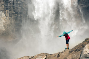 A huge waterfall in the background with a female hiker in a hooded blue and red jacket, black tights looking down at her feet balancing on a rock in the right side of the photo.