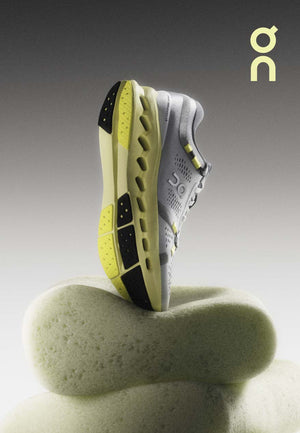 Vertical picture of the On Cloudsurfer 2 toe down on foam with a neutral grey background.  The shoe is light grey with a yellow sole.  The On logo is in yellow and in the top right corner