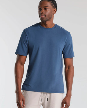 Man wearing a blue t-shirt with a white brand logo on a light gray background