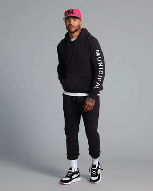 Man wearing a black hoodie with 'Municipal' text, black pants, and a pink cap on a gray background