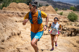 Two runners in trail gear running a trail.  The first runner is a male in focus wearing blue and orange apparel and the second runner is a female slightly out of focus wearing a white top and orange shorts.  Both are wearing sunglasses