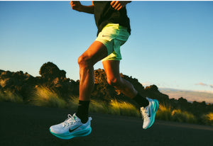 Image of a male runner from the waist down wearing yellow shorts and white Nike sneakers on a grassy field with trees and blue sky as the background.